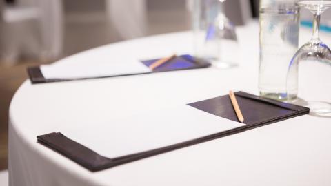 A notepad, pen, and empty glass are set on a table covered in a white tablecloth.