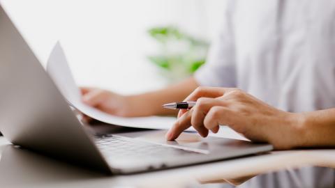 A person sits behind a laptop, holds a pen and is typing on the keyboard.