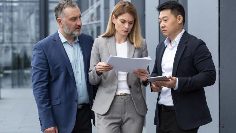 Three adults stand outside an office building wearing suits and looking at a set of papers.