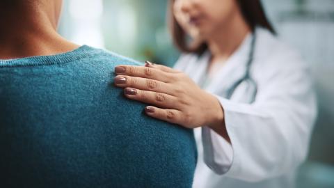 A woman wearing a white doctor's coat rests her hand on a patient's back to show support.