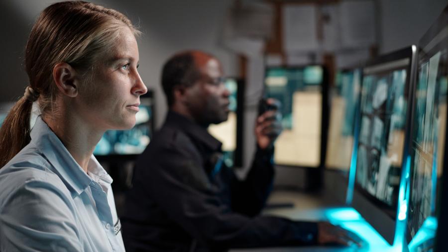 Two people sit at a desk and are looking at computer monitors of security camera footage.