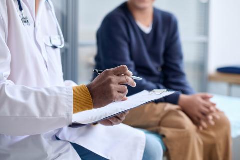 A doctor holds a clipboard and a pen and a young child sits on an examination table.