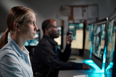 Two people sit at a desk and are looking at computer monitors of security camera footage.