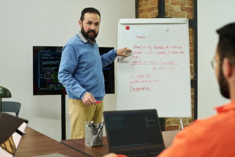 Middle aged man teaching programming concepts on whiteboard to adult male student in classroom setting, discussing code structure with laptops and digital screen visible.